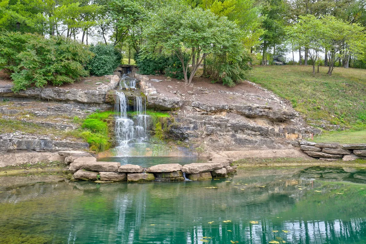 A tranquil waterfall flows into a reflective pond, surrounded by lush greenery and rock formations. The area exudes peace and natural beauty, perfect for nature walks, picnicking, or quiet reflection.