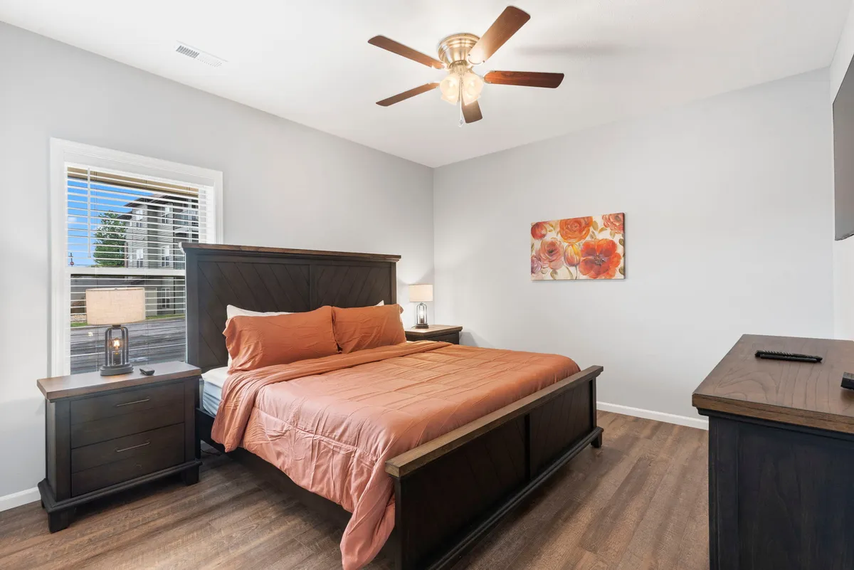 A warmly decorated king bedroom with coral bedding, wooden nightstands, colorful wall art, and a ceiling fan. Sunlight filters through the window, creating an inviting and restful atmosphere.