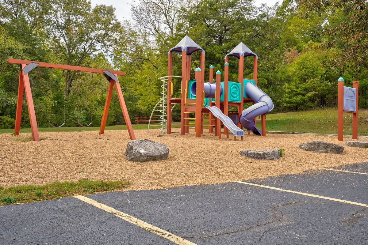 A cheerful playground scene featuring colorful play structures, swings, and rock features, with children and families enjoying outdoor activities together in a safe, natural environment.