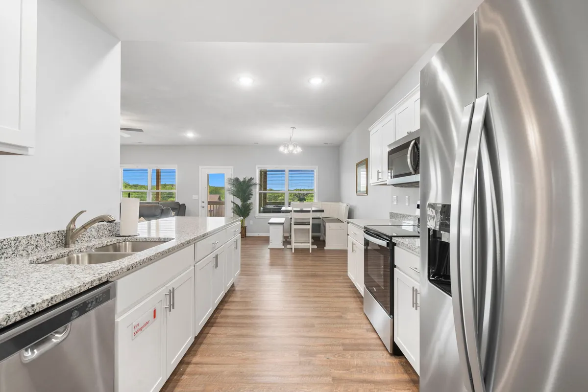 A wide view of a welcoming, modern open-concept space featuring a kitchen with granite countertops and stainless steel appliances, a dining table near large windows letting in ample natural light, and a seamless flow between kitchen and dining areas.