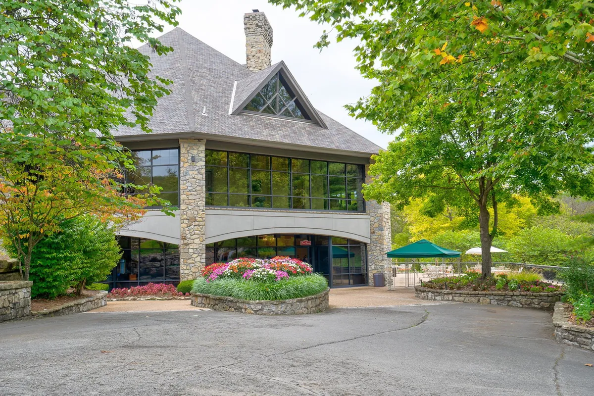 A welcoming exterior scene featuring large glass windows, a stone facade, and vibrant flower beds surrounded by trees. The building projects openness and community, connecting indoor amenities with the natural world.