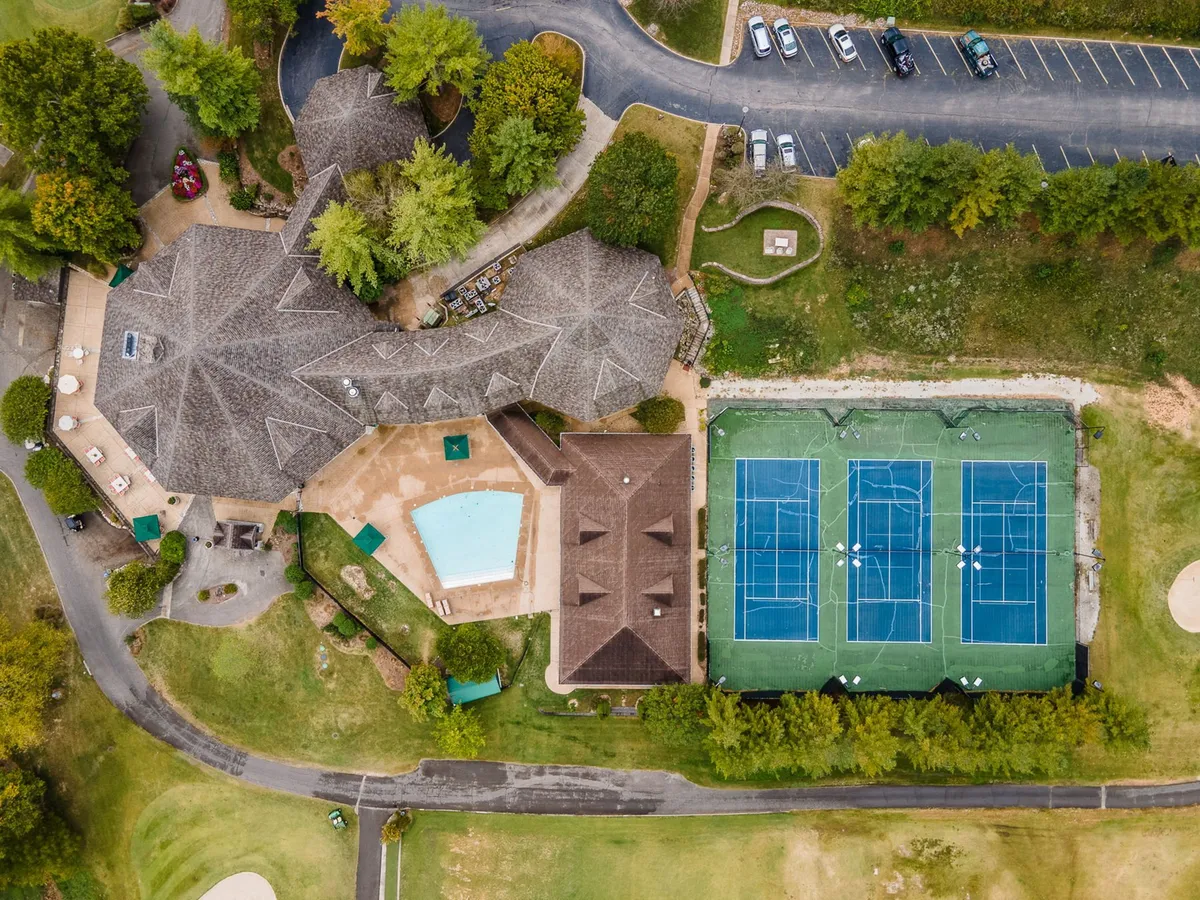 An exterior view showing the community center, swimming pool, tennis courts, and beautifully landscaped gardens. The lively, active mood captures families and groups engaging in swimming, tennis, and social gatherings outdoors.