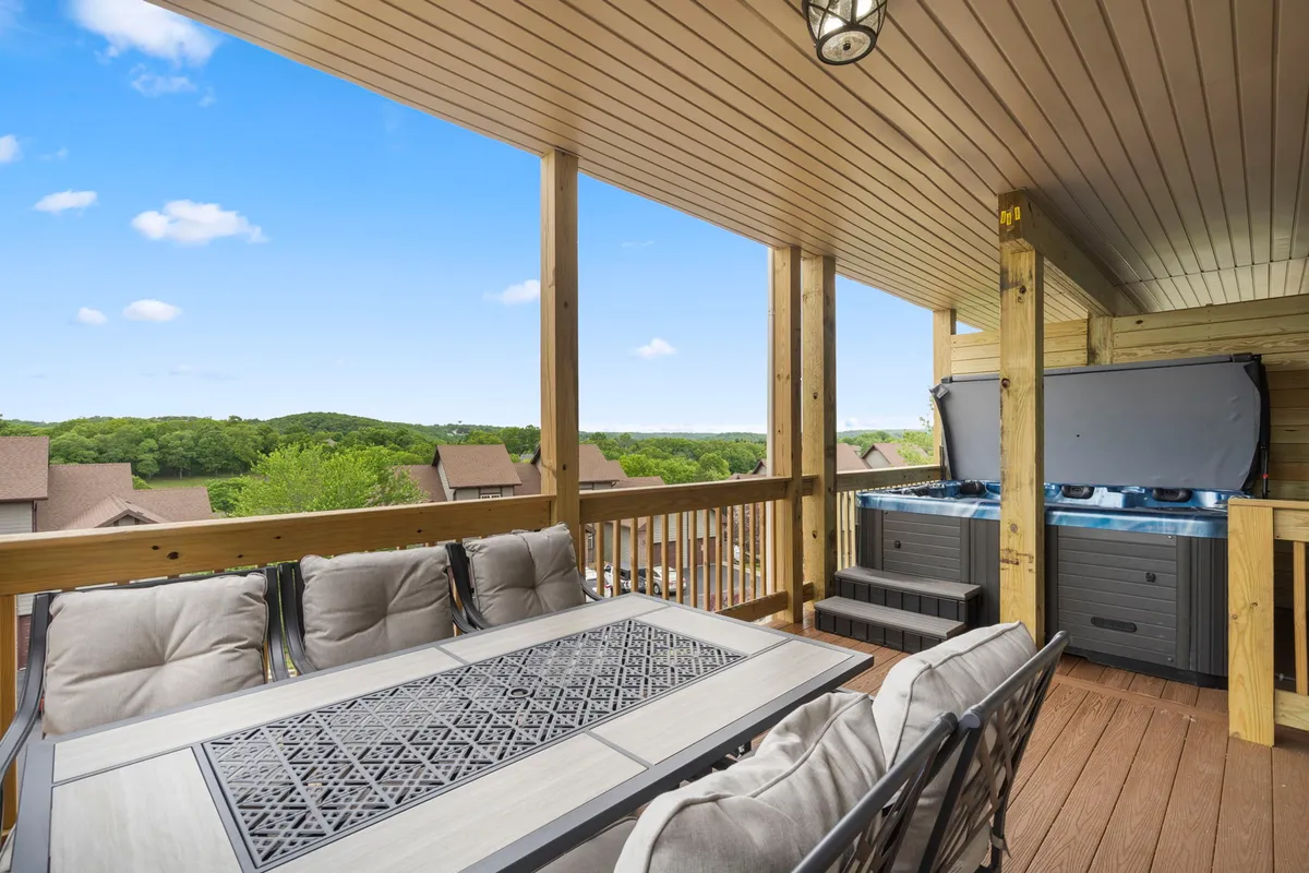 A spacious private deck featuring an outdoor dining table, inviting seating, a hot tub, and views of the surrounding landscape. The setting is tranquil, with natural greenery and hills visible in the background, suggesting both relaxation and a connection to the resort scenery.