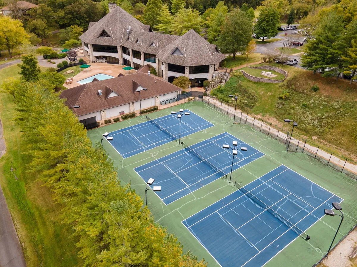 The image features tennis courts adjacent to a clubhouse, framed by mature trees and with the pool area visible in the background. The scene exudes activity, with spaces designed for friendly tennis matches, social events, and swimming.